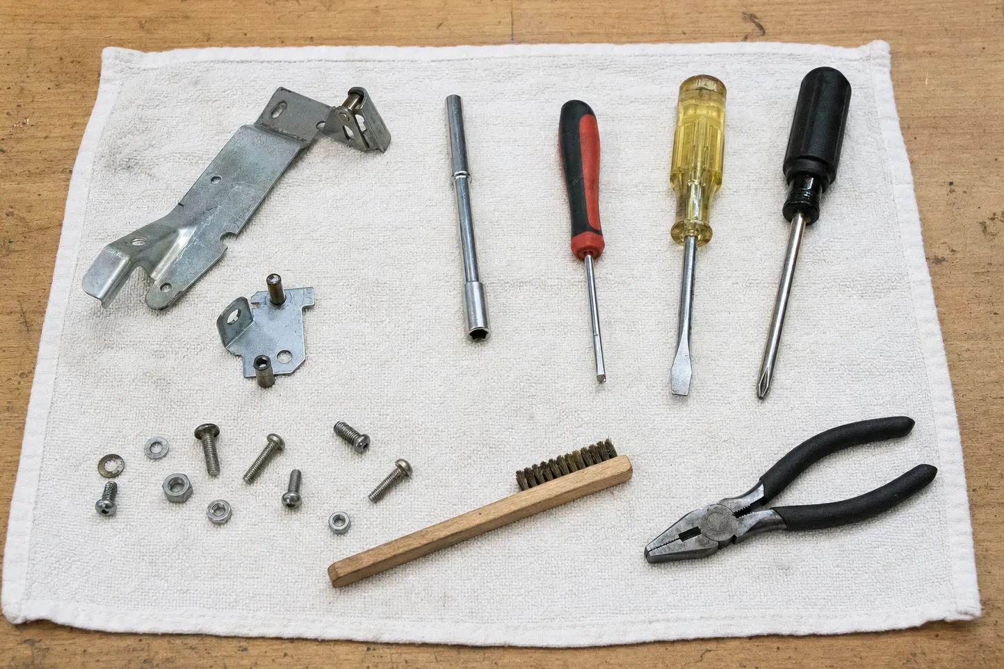 Oven door hinge arm and a latch bracket beside screws, nuts, a nut driver and pliers on a white towel